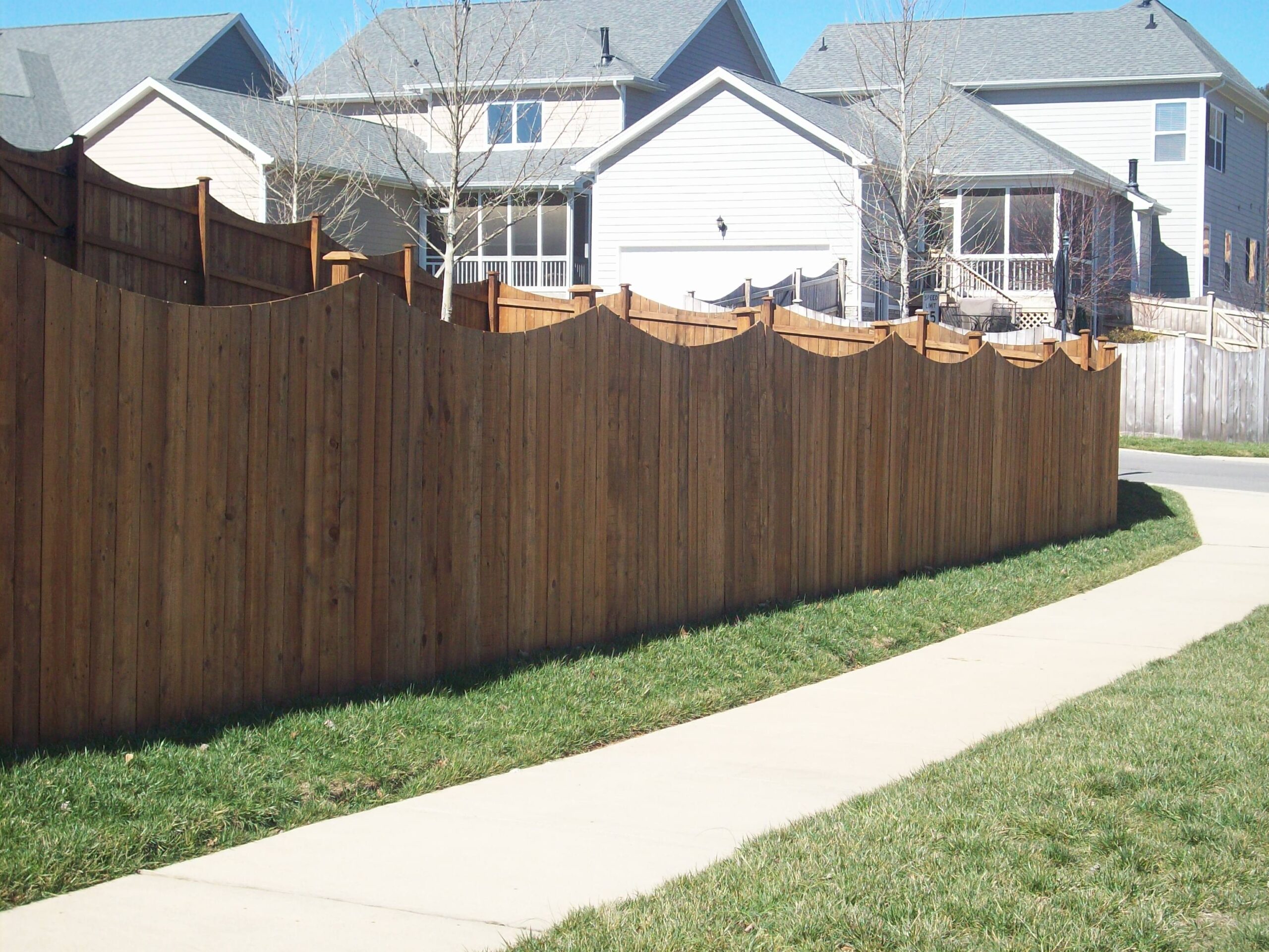 Wooden fence with a wavy top design, well-maintained along a sidewalk, showcasing clean and sturdy posts, ideal for residential areas. Regular cleaning and sealing can preserve its appearance and longevity.