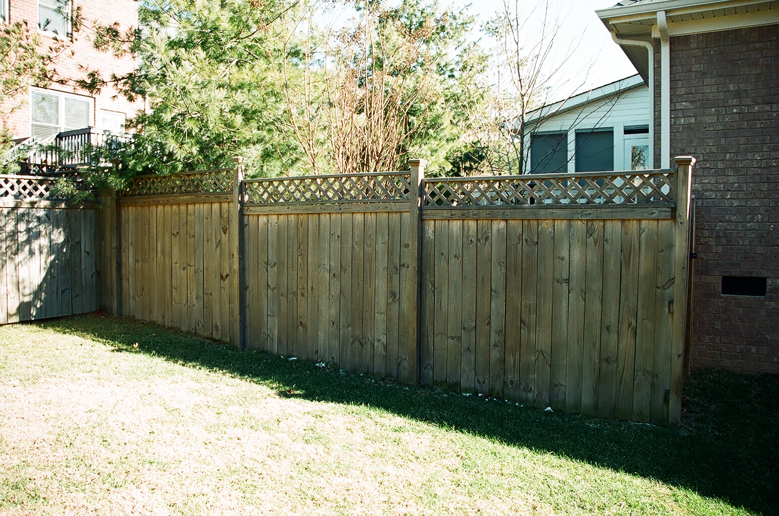 Wooden fence with a decorative lattice top, running along a grassy area with trees and a house visible in the background.